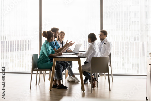 Five multi-ethnic medical workers take part in scheduled meeting in modern clinic conference room, sit at table and lead talk, share opinions, professional vision and skills. Collaboration, medicine