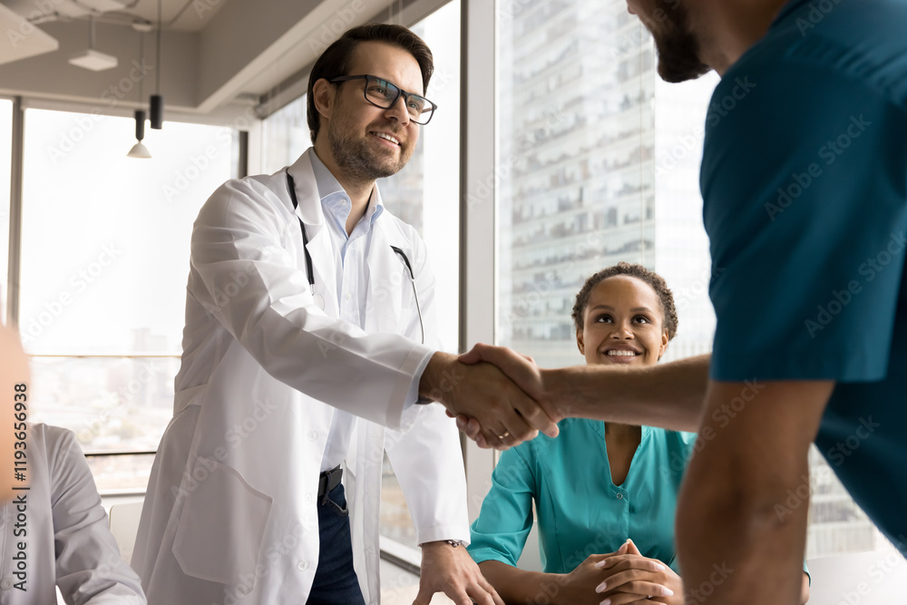 Obraz premium Two male doctors in uniforms shaking hands, start medical concilium in clinic office boardroom with diverse staff members. Gesture of mutual respect, agreement, or conclusion of successful discussion