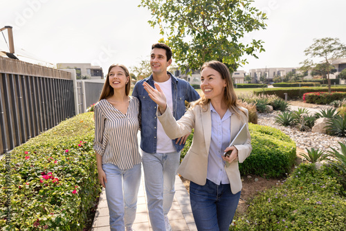 Happy professional realtor woman giving tour around well suburb neighborhood for house buyers, showing apartment exterior, location to young couple of customers, walking outside, pointing hand away