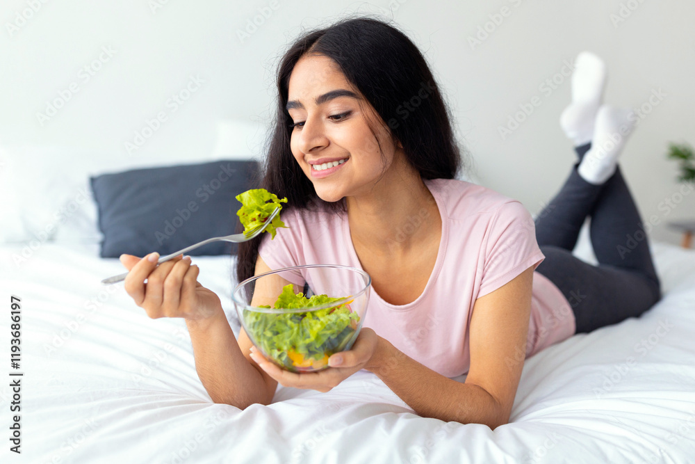 Balanced raw food diet. Lovely Indian woman eating fresh vegetable salad, lying on bed at home with yummy meal. Positive Hindu lady having vegan lunch, taking care of her wellbeing Balanced raw food diet. Lovely Indian woman eating fresh vegetable salad, lying on bed at home with yummy meal. Positive Hindu lady having vegan lunch, taking care of her wellbeing