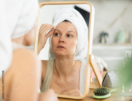 Careful middle-aged lady viewing her complexion in a spacious mirror while seated at a kitchen-table in her house photo