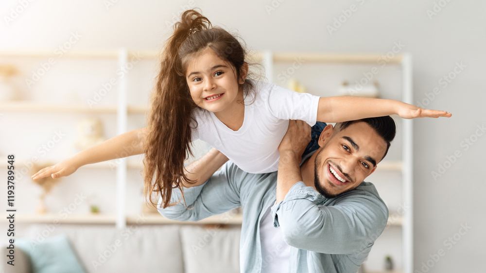 Fototapeta premium Happy arab father and daughter having fun together at home, middle-eastern young man dad playing airplane with his cute daughter in living room, lifting kid up, holding on shoulder, copy space