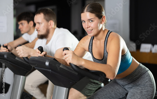 Girl hardly perform track on stationary bicycle, pedaling and exercising in fitness class. Fitness equipment, athlete engaged in physical task during training, work out, participate in sport challenge photo