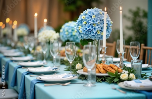 Beautifully decorated table setting for wedding baby shower party. Blue flowers in centerpiece arrangement alongside white candles. Table set with plates, glasses, napkins. Small pastries placed on