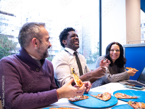 Three colleagues laugh heartily while enjoying pizza around a table with a laptop and phone, set against a bright city backdrop, embodying teamwork and a positive workplace vibe
