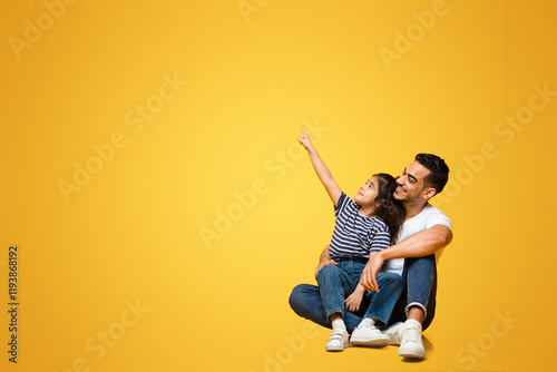 Cheerful arab dad and little daughter pointing aside at copy space over yellow background, positive middle eastern father and child sitting on floor and looking at free place with interest photo