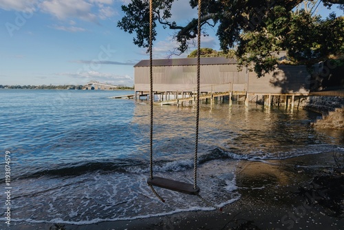 Empty swing by the water, by a wooden pier, with a bridge in the background. Tranquil coastal scene. Herne Bay, Auckland, New Zealand photo