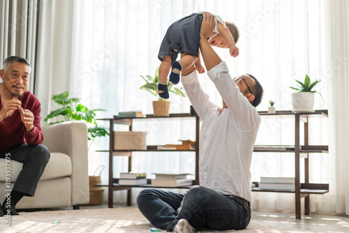 Joyful moment of multigenerational family at home, father holding up his baby smiling and playing while the grandfather looks on with a big smile enjoying the happy interaction, love within a family photo