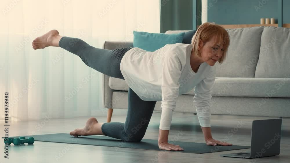 A senior woman is engaging in a home workout, focusing on her fitness routine. She maintains balance on a mat, using a laptop for guidance while exercising in a bright living space.
