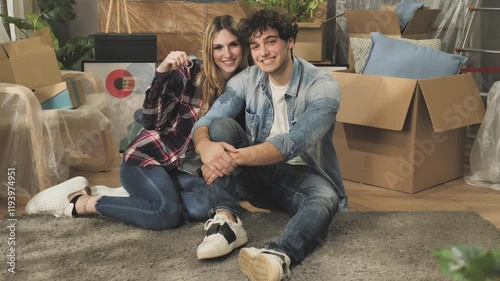happy young caucasian couple sits on the floor of new apartment fill of boxes with their belongings showing house key,people moving to new rental home 
