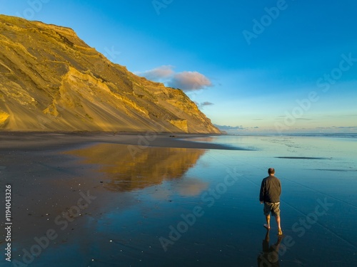 Man barefoot walks along a reflective beach at sunset, coastal cliffs in the background. Tranquil scene. Hamiltons Gap, Awhitu, Auckland, New Zealand photo