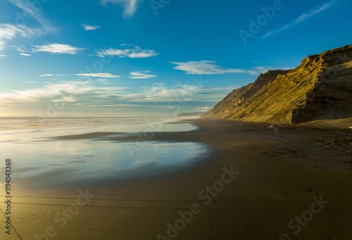 Golden hour beach scene; calm waves, dark sand, and dramatic cliffs. Few people on the shore. Hamiltons Gap, Awhitu, Auckland, New Zealand photo