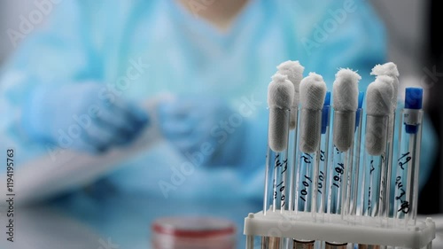 Close-up of test tubes with images of tests in the laboratory. In the background, a doctor in a blue laboratory suit opens a swab package.