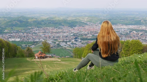 A young girl sitting on a scenic hilltop at Pohorje, overlooking the city of Maribor. The landscape features a panoramic view of the urban skyline, lush forests, and a serene atmosphere. photo