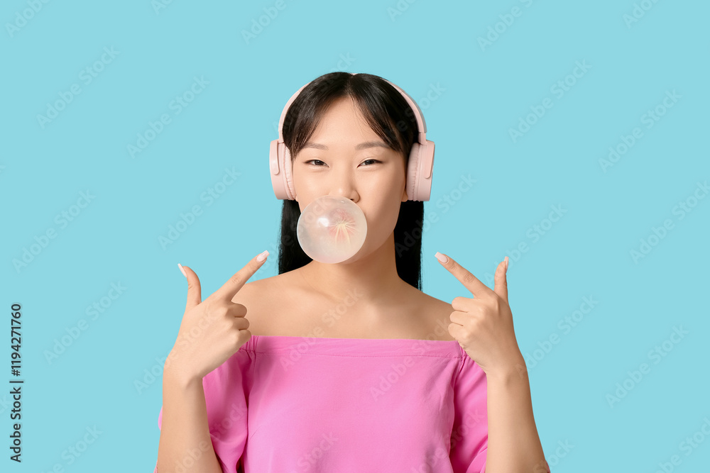Pretty young Asian woman in headphones blowing bubble gum and pointing at it on blue background Pretty young Asian woman in headphones blowing bubble gum and pointing at it on blue background