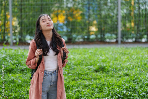 Young asian student is enjoying the fresh air in a university campus park, holding her backpack straps and closing her eyes, surrounded by lush greenery