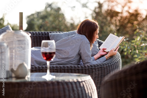 A woman is relaxing on the veranda of a country house and reading a book over a glass of red alcoholic drink or wine. The concept of relaxation and luxury wine.