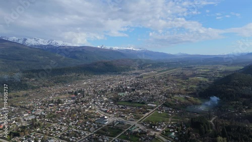 High-resolution aerial drone footage of El Bolson nestled in a scenic valley, surrounded by majestic mountains, forests, and green fields under a partly cloudy sky. photo