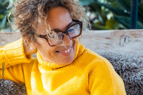 Positive lifestyle portrait of a mature woman with curly hair, glasses, and a bright yellow sweater, sitting outdoors in her garden, smiling as she enjoys the peacefulness and natural tranquility photo