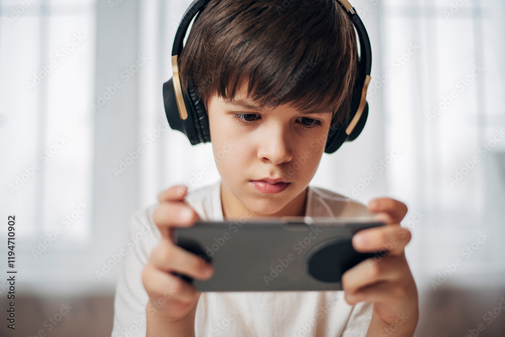 Cute boy with headphones playing games on his phone at home. Happy child looking at his smartphone. Cute boy with headphones playing games on his phone at home. Happy child looking at his smartphone.