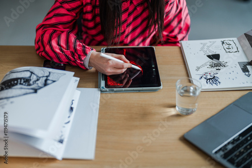 digital artist concentrates on creating intricate artwork using a stylus and tablet, seated at a well-equipped desk with sketchbooks, water, and art supplies in a modern workspace. photo