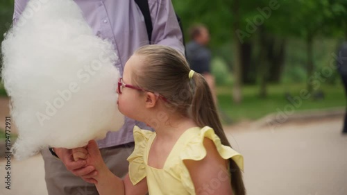 Girl with Down syndrome wearing pink glasses and yellow dress excitedly reaches for cotton candy held by woman in purple shirt during walk in park. Scene symbolizes joy and carefree moment photo