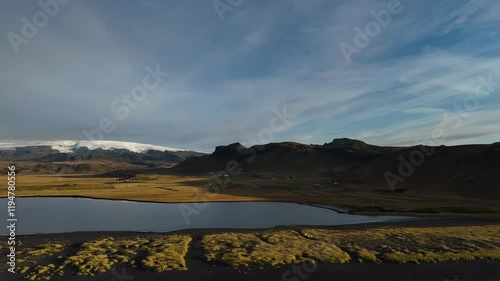 A breathtaking aerial view of Iceland’s rugged terrain, featuring serene lakes, golden grasslands, and majestic snow-capped mountains in the distance.