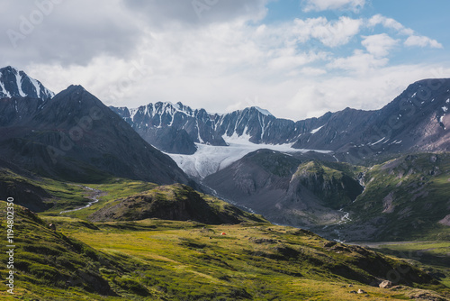 Foto Dramatic scenery in alpine valley with creek among green hills and rocks with view to rocky pointy peak, large snow-capped peaked top, mountain range and big glacier tongue under clouds in blue sky