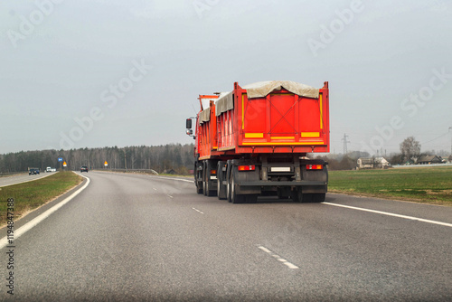 An orange grain dump truck with a semi-trailer transports grain along the road from a farm. Transportation of grain harvest. Copy space for text, agricultural photo