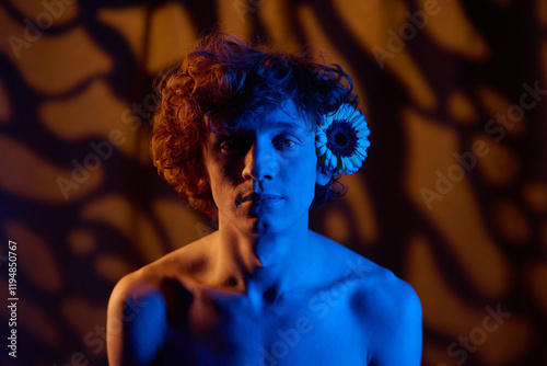 Sad young adult man with flower behind ear sitting still looking at camera and illumed with deep blue neon light photo