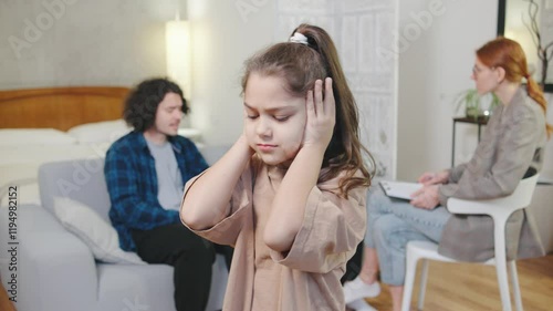 In a cozy, bright apartment, a girl holds her head and covers her ears while her parents argue in the background. The young psychologist tries to help, reflecting the intense emotional tension
