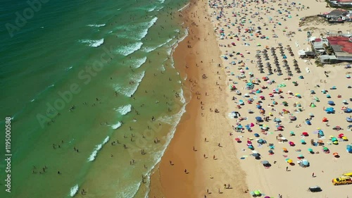 Aerial view busy beach filled with swimmers and sunbathers under vibrant umbrellas. Waves crash gently on the shore as families enjoy the warm summer atmosphere.