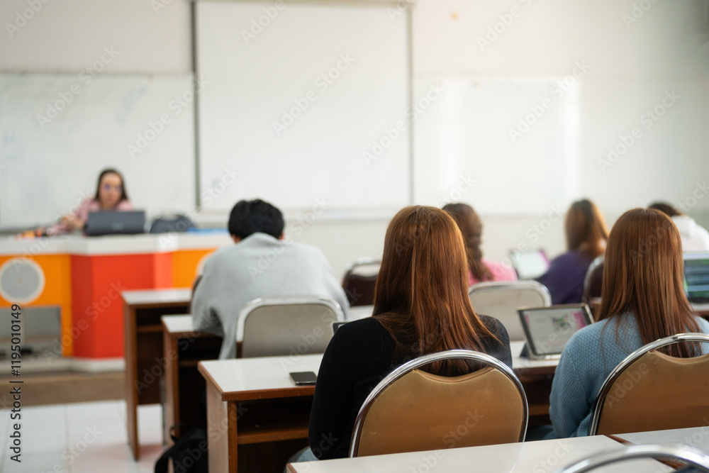 Students engage in a lecture, sitting at desks with their attention focused on the lecturer at the front of the classroom. The lecturer uses a projector screen for the presentation. Students engage in a lecture, sitting at desks with their attention focused on the lecturer at the front of the classroom. The lecturer uses a projector screen for the presentation.