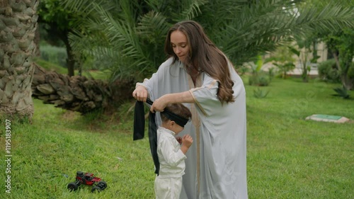 Mother ties blindfold over her son's eyes, preparing for a playful game of tag in lush garden of family estate. With a smile, she quickly steps away, adding excitement and joy to their bonding moment