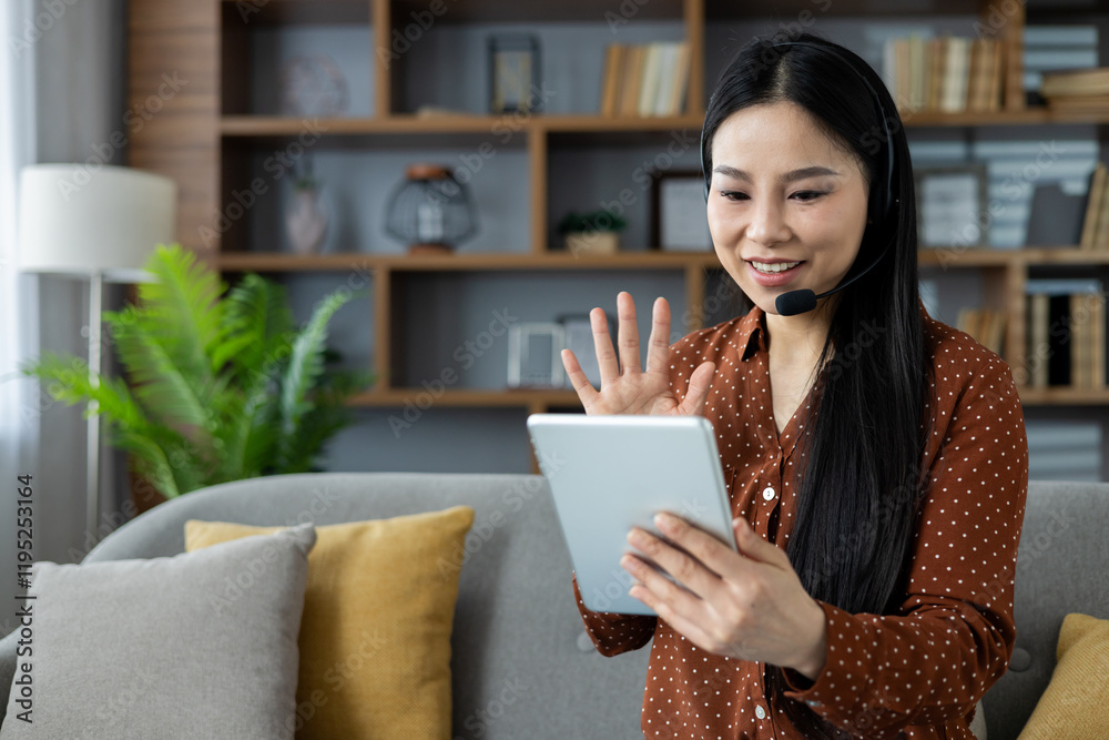 Asian woman participates in virtual meeting at home using tablet and headset. She is sitting on sofa, waving and smiling, demonstrating technology's role in remote communication.