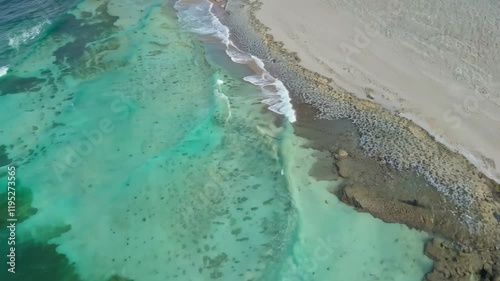 Aerial view of a clear turquoise ocean meeting a sandy shore with rocky formations.