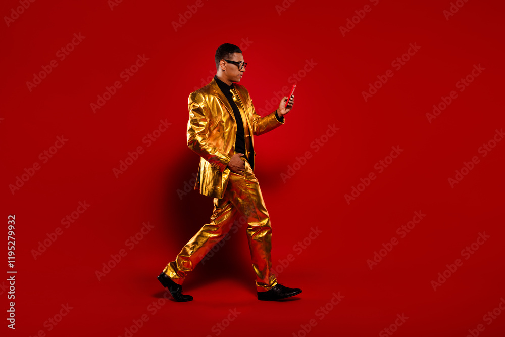 Charismatic man in glossy golden suit walks elegantly with phone against vibrant red background Charismatic man in glossy golden suit walks elegantly with phone against vibrant red background