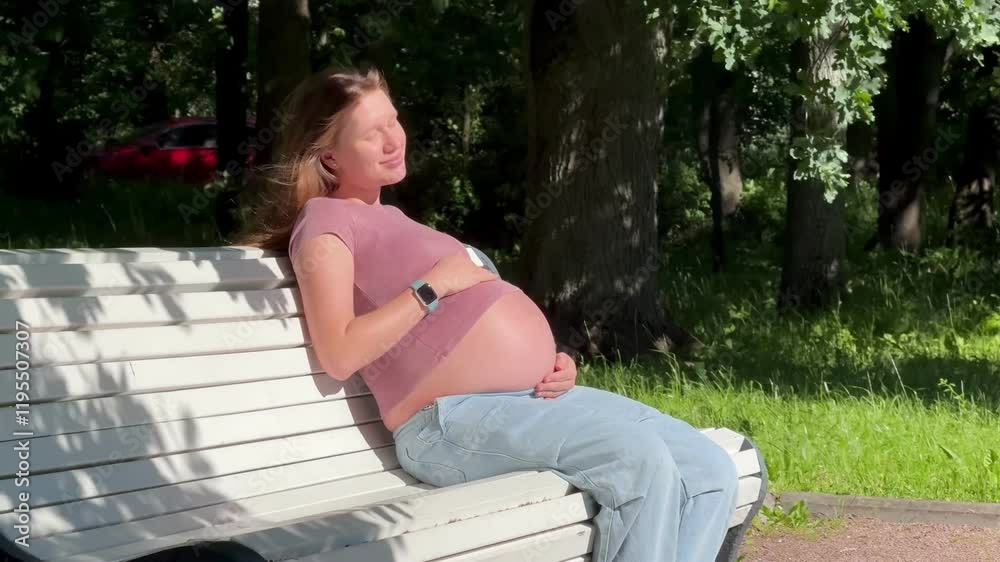 Young happy pregnant woman is walking, sitting on bench in the summer park and stroking her belly