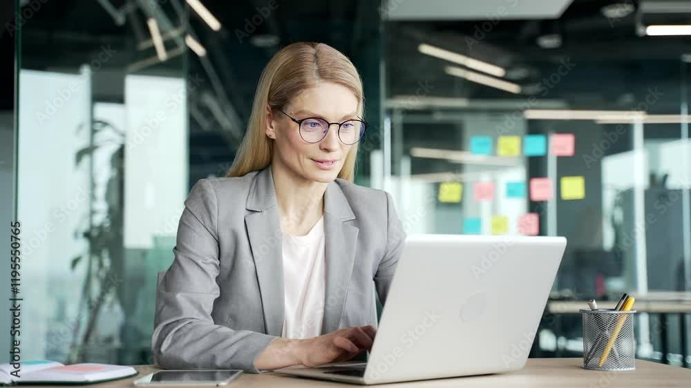 Confident businesswoman in a jacket typing on laptop sitting at workplace in business office. Female employee in glasses working in computer application, banking, texting a client or chatting online