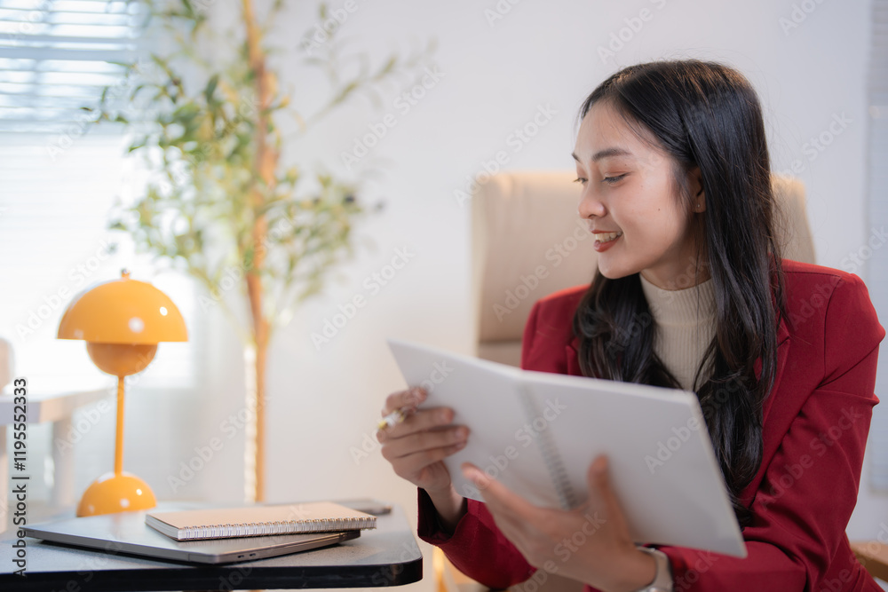 Young Asian businesswoman wearing red suit reading notes while sitting comfortably in her office with laptop, notepad and smartphone on the desk Young Asian businesswoman wearing red suit reading notes while sitting comfortably in her office with laptop, notepad and smartphone on the desk