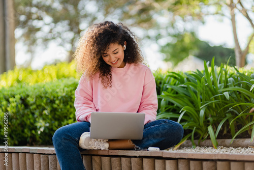 pretty curly young woman sitting in park working on laptop remote job