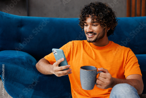 Young Indian man wear orange casual clothes sit near blue sofa couch hold use mobile cell phone drink tea stay at home hotel flat rest relax spend free spare time in living room indoor Lounge concept photo