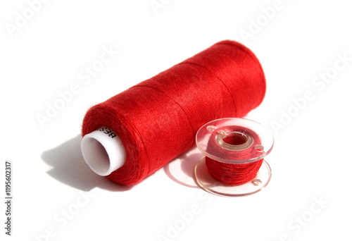 On a white background lies a red spool of thread and a bobbin.	