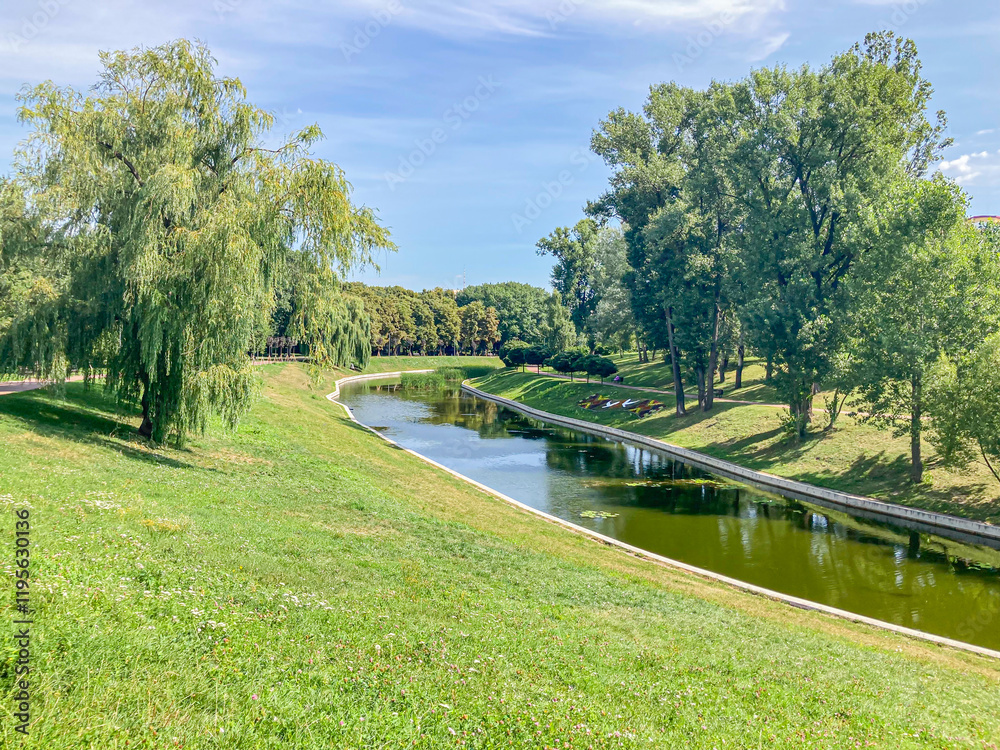 A serene park scene showcases a curved path, small riverpond, lush green grass, well-maintained ground, and various tree species against a clear blue sky A serene park scene showcases a curved path, small riverpond, lush green grass, well-maintained ground, and various tree species against a clear blue sky