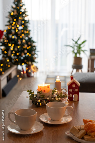 Wallpaper Mural Cozy Christmas scene with two empty white cups on wooden table, festive candles, and plate of snacks in the foreground, with decorated Christmas tree glowing in soft bokeh lights in the background. Torontodigital.ca