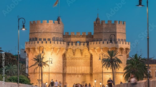 Serrans Towers (Torres de Serranos) day to night transition timelapse in Valencia, Spain. A grand medieval gate from city's ancient walls. People walking on a bridge near liiuminated historic landmark photo