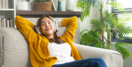 Attractive young Asian woman leans back on sofa, wearing headphones and smiling, enjoying a weekend with positive music. Smiling Asian woman listening to music relaxing carefree digital lifestyle.