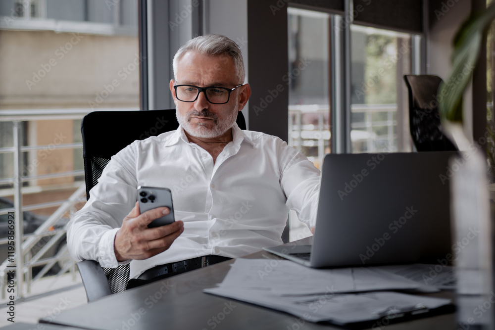A mature businessman with glasses sits at his office desk, holding a phone, checking emails or reading messages. Focused, professional, and engaged in work. A mature businessman with glasses sits at his office desk, holding a phone, checking emails or reading messages. Focused, professional, and engaged in work.