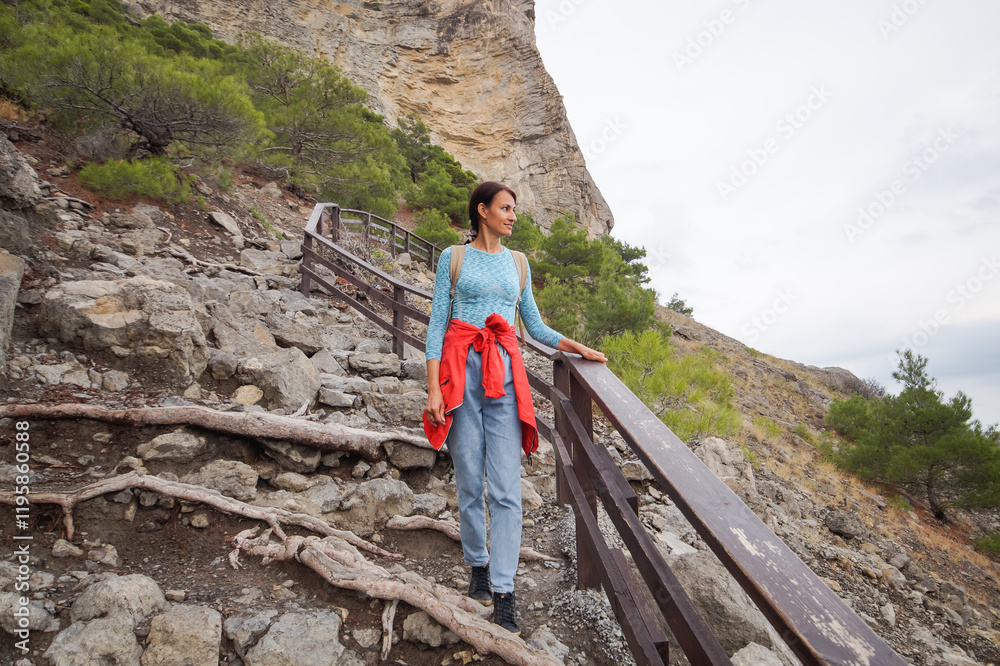 Happy female tourist with a backpack on the top of a mountain, enjoying the sea view, standing on a rock and looking into the distance. The village of Novy Svet in Crimea. Russia