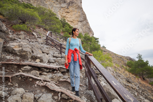 Happy female tourist with a backpack on the top of a mountain, enjoying the sea view, standing on a rock and looking into the distance. The village of Novy Svet in Crimea. Russia